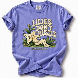 Lilies Don’t Hustle Premium T-Shirt in Flo Blue, front view with botanical character inspired by Matthew 6:28–29
